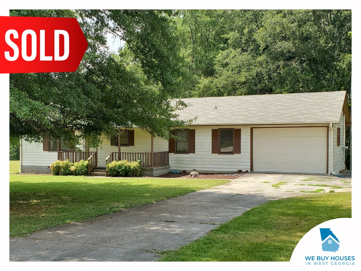 A ranch-style house with white siding, brown trim, and large tree in the front yard, bought by We Buy Houses in West Georgia